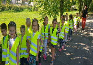 dzieci ubrane w kamizelki odblaskowe w kolorze cytrynowym idą parami po chodniku, jego prawą stroną pod opieką nauczycielki